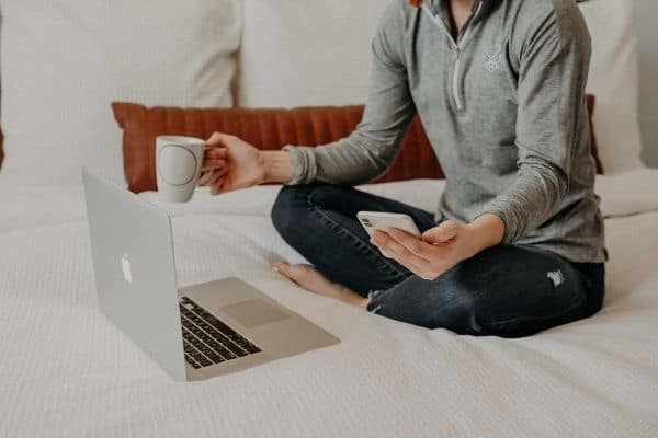 woman holding a cup of coffee in one hand, her phone in the other and a laptop in front of her on the bed
